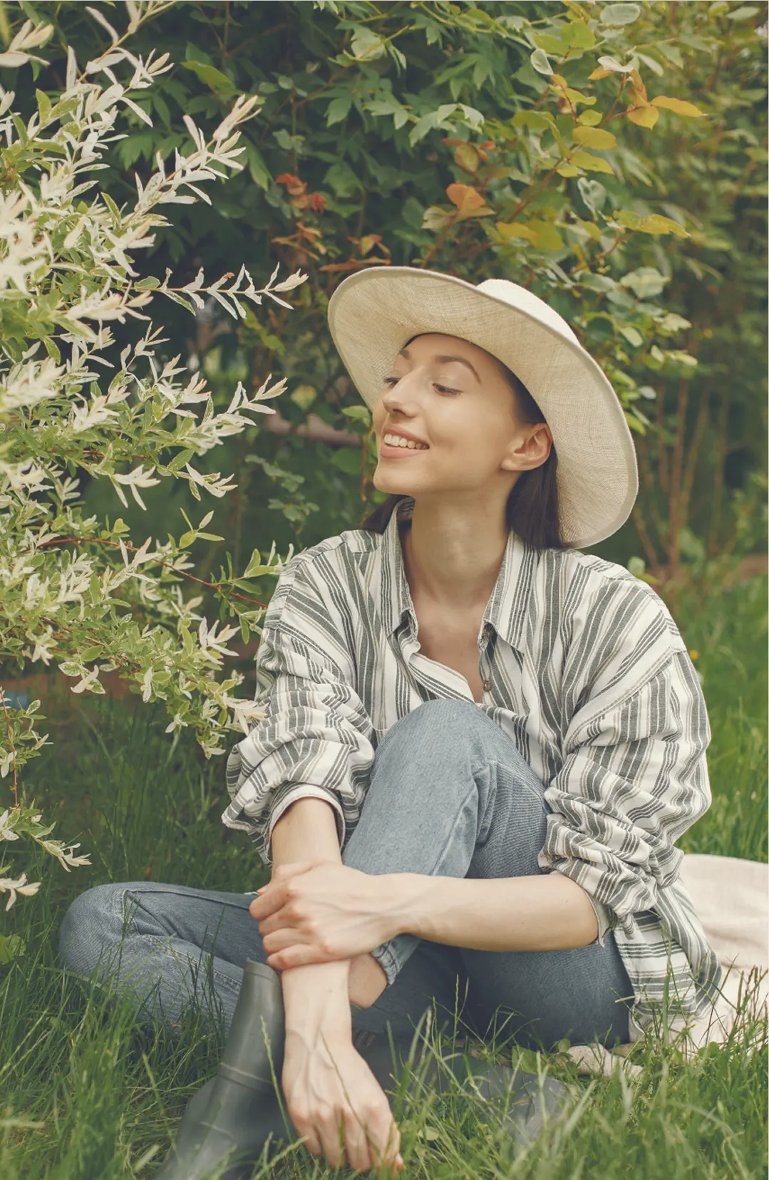A woman in a sun hat sitting in the grass beside a flowering shrub, unhurried and at ease.