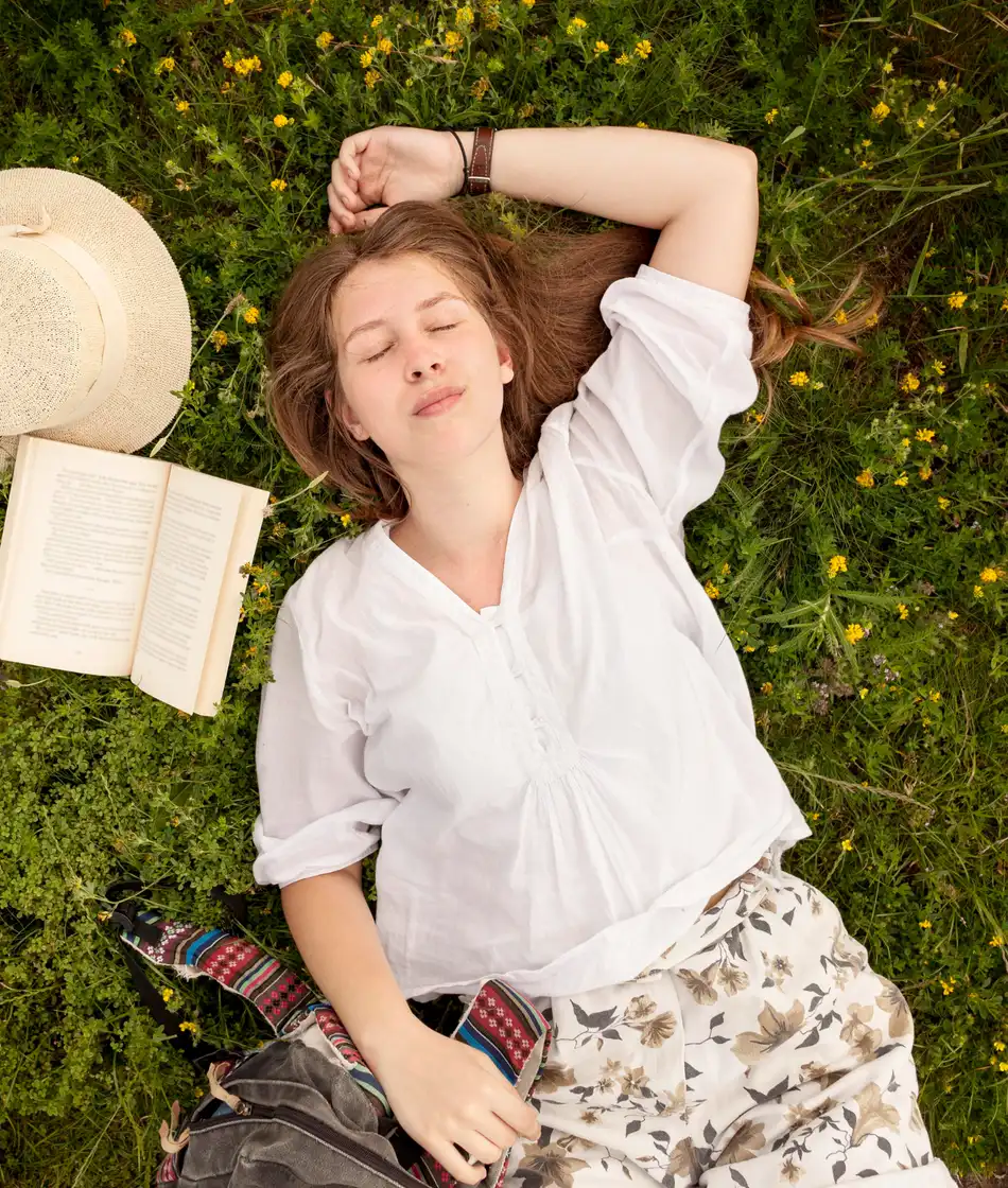 A woman lying in a wildflower meadow next to an open book and a straw hat, resting between tasks.